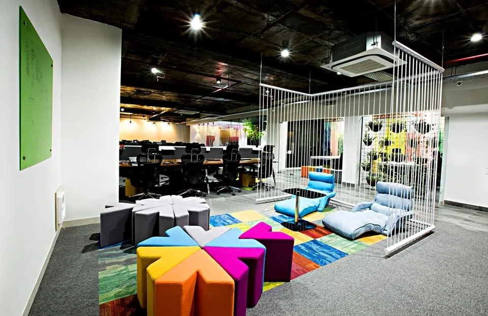 Breakout lounge with colourful modular seating on patterned carpet, beside a white rod screen with hanging planters