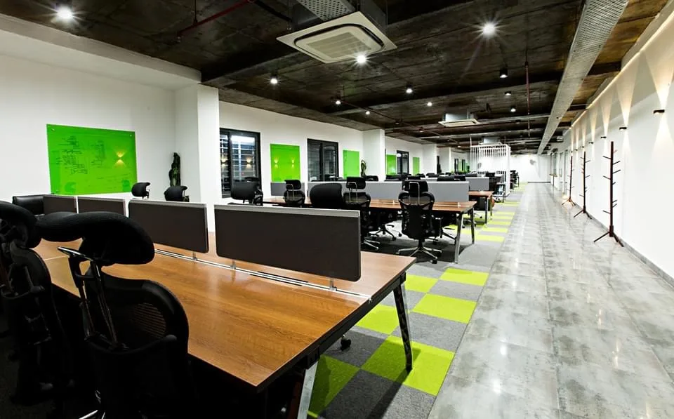 Rows of open workstations with timber desks and grey screens, green glass writing boards and an exposed dark ceiling with cassette air conditioning units